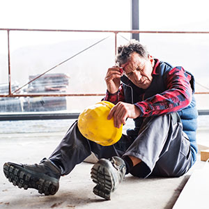 A distressed construction worker holding a hard hat and sitting on the floor, representing a workplace injury.